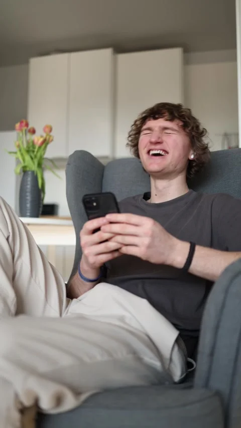 Vertical screen: Young man laughing uncontrollably while looking at smartphone,  Stock Footage 302632204