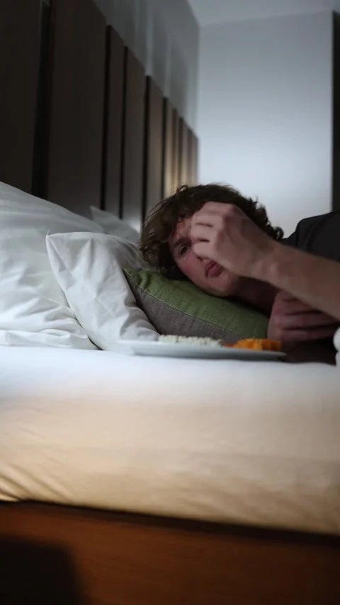 Vertical screen young man lies in bed late at night picking food from a plate wi Video stock 318217541