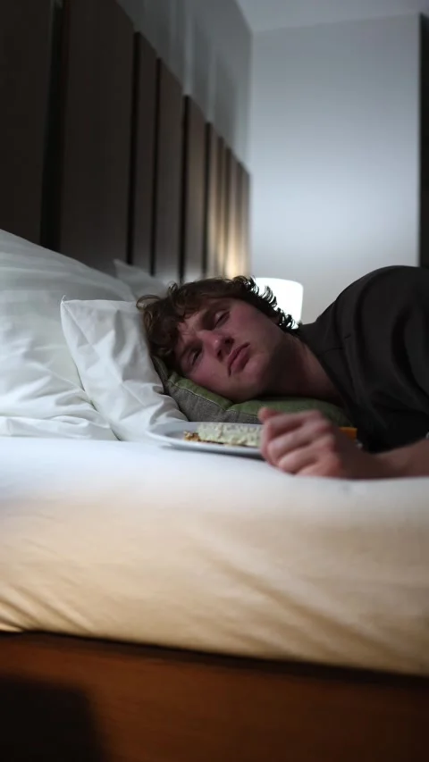 Vertical screen young man lies in bed falling asleep with food on a plate nearby Stock Footage 318217545