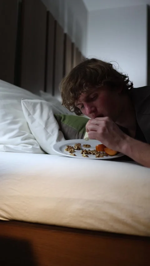 Vertical screen young man lies in bed late at night binge eating cheese and nuts Stock Footage 318217558