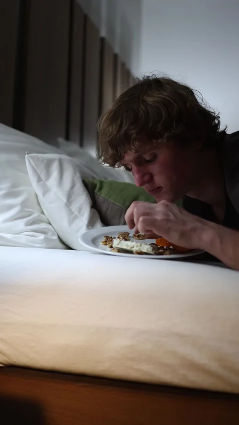 Vertical screen young man lies in bed late at night chewing on cheese and nuts s Stock Footage 318217559