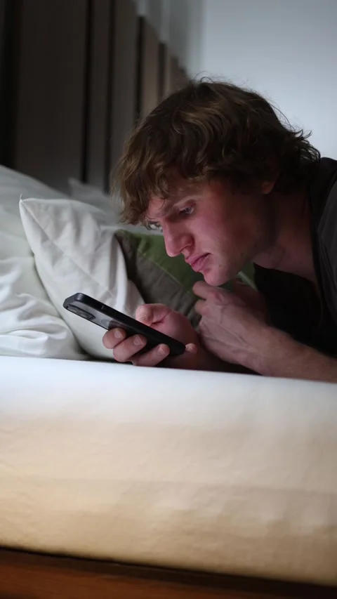 Vertical screen young man lies in bed focused on his phone late at night scrolli Stock Footage 318217587