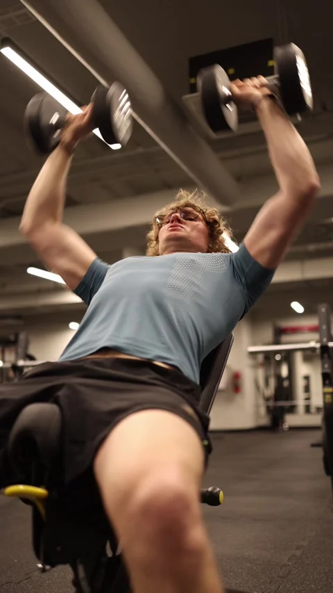 Vertical screen: A young man is lifting dumbbells on a bench press, focusing on  Stock Footage 301676871