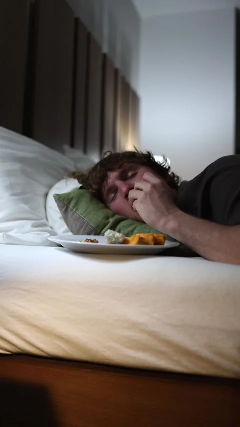 Vertical screen young man lying on bed late at night eating snacks from plate un Stock Footage 318217644