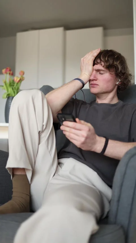 Vertical screen: A young man sits on a couch, holding his smartphone with a dist Stock Footage 302632544