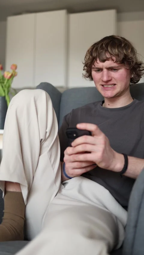 Vertical screen: A young man sits on a couch, holding his smartphone with an awk Stock Footage 302632549
