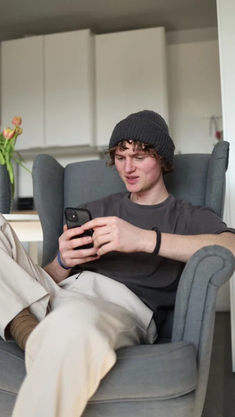 Vertical screen: A young man sits comfortably in a chair, smiling while using hi Stock Footage 302633251