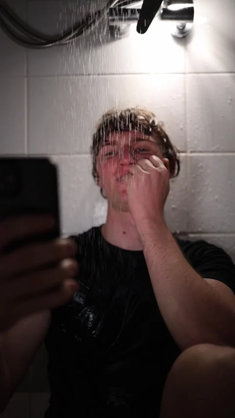 Vertical screen young man sits under shower holding smartphone while laughing at Stock Footage 316066148