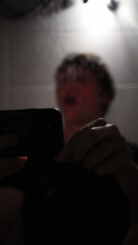 Vertical screen young man sits under shower water falling as he holds smartphone Stock Footage 316066177