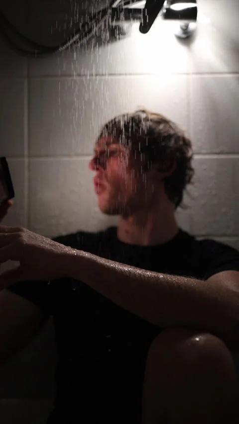 Vertical screen young man sits under shower streams holding smartphone watching  Stock Footage 316066269