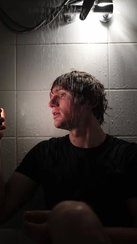 Vertical screen young man sits under running shower holding smartphone as water  Stock Footage 316066305