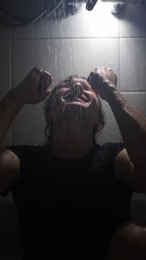 Vertical screen young man sits under shower raising clenched fists to face water Stock Footage 316066345