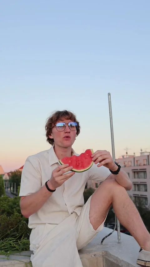Vertical screen young man in summer outfit enjoying fresh watermelon at sunset s Stock Footage 315413747