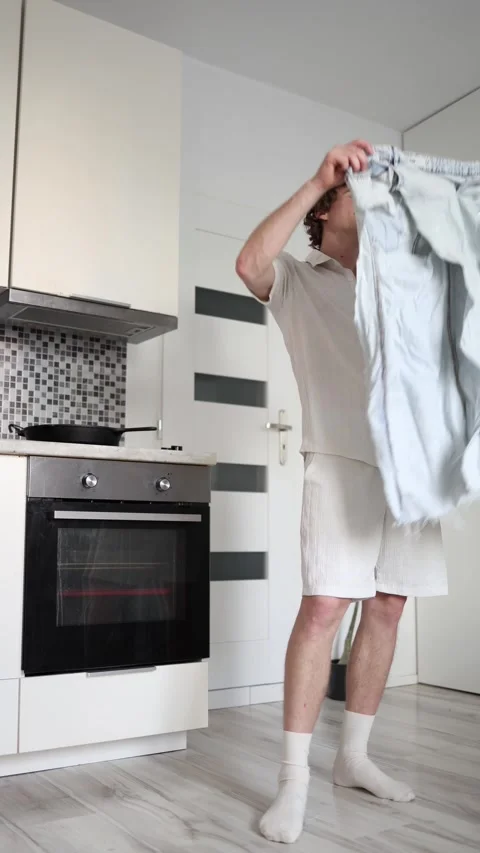 Vertical screen young man trying on oversized ripped jeans in bright kitchen loo Stock Footage 318867726