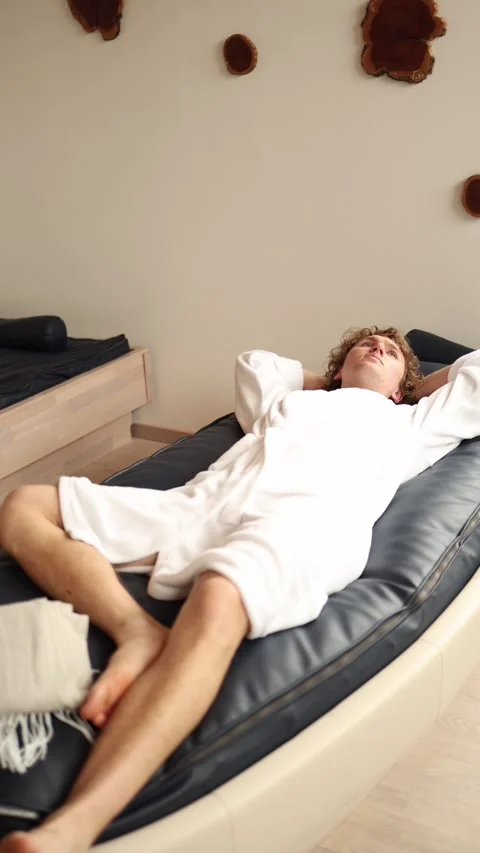 Vertical screen young man in white bathrobe lies on modern waterbed with closed  Stock Footage 318217427