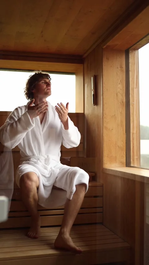 Vertical screen young man in white bathrobe sits inside wooden sauna raising han Stock Footage 318217450