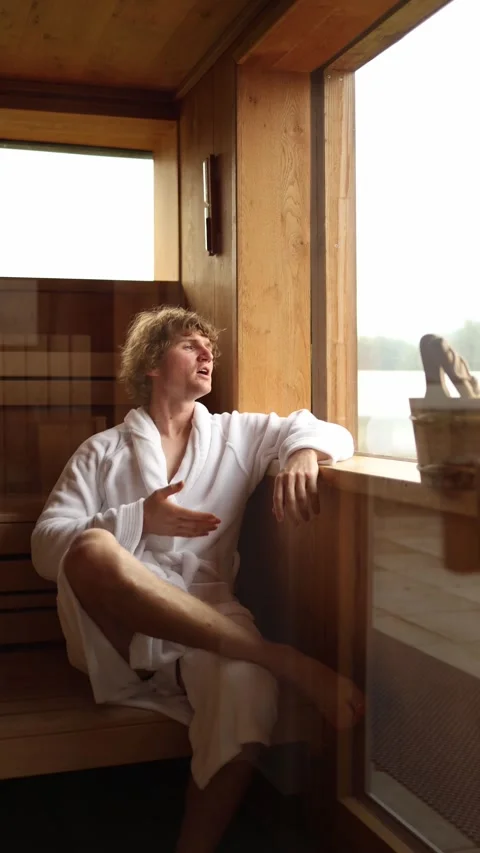 Vertical screen young man in white bathrobe relaxing inside wooden sauna near wi Video stock 318217461