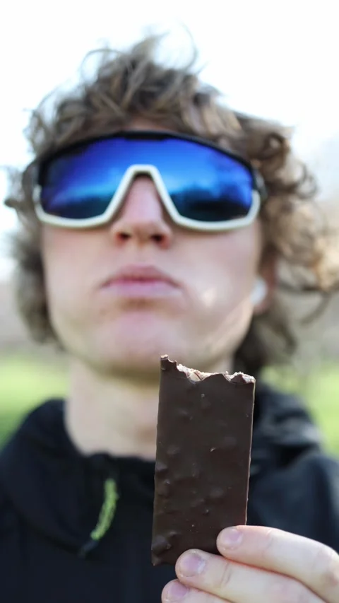 Vertical screen. Young person in reflective sports sunglasses biting into a choc Stock Footage 305999148