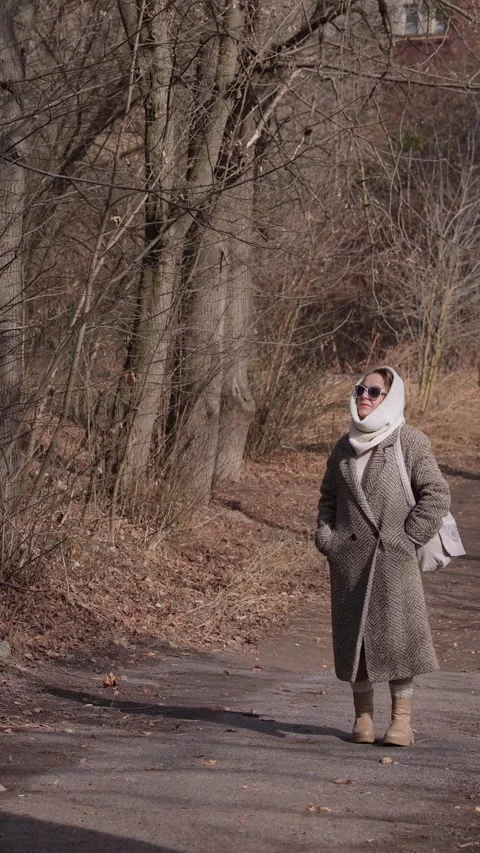 Vertical. Senior lady in coat with woolen head-wrap walks along road among trees Video stock 329465976