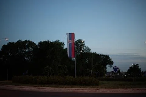 Vertical Serbian flag hangs from a flagpole in a public roundabout at dusk Stock Photos