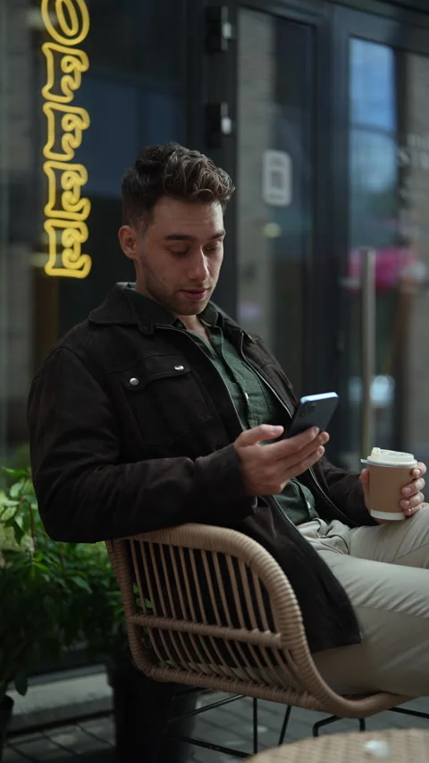 Vertical shot of handsome young man sitting at outdoor cafe chair, holding Stock Footage 297630980