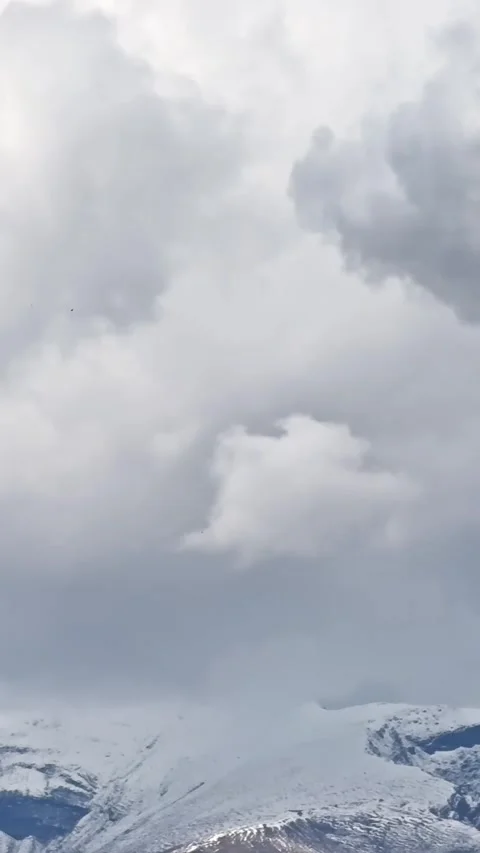 Vertical shot of heavy grey clouds moving above snow-covered mountain peaks Видео 339075939