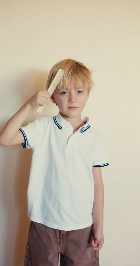 A vertical shot of a six-year-old blond boy in a white T-shirt combing his hair Video stock 323107219