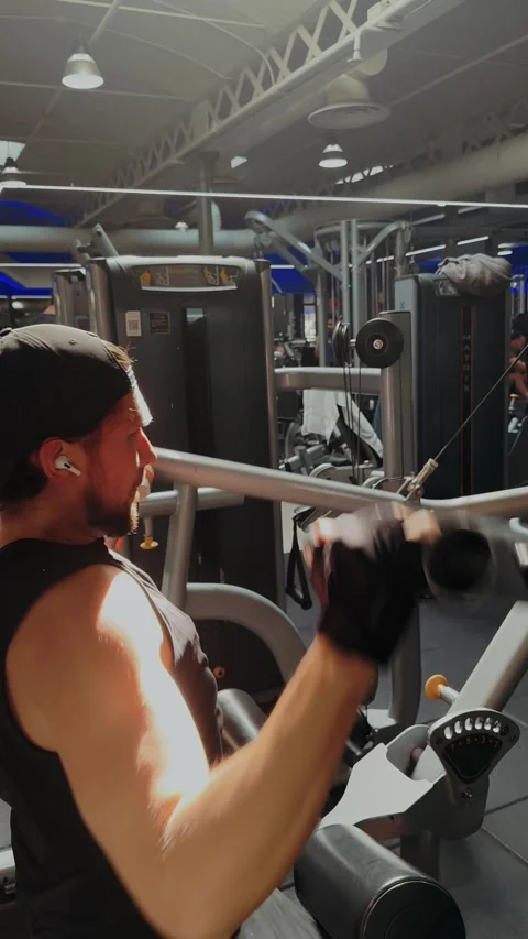 Vertical shot of a young man exercising on lat pulldown machine in a bright gym Stock Footage 334719555