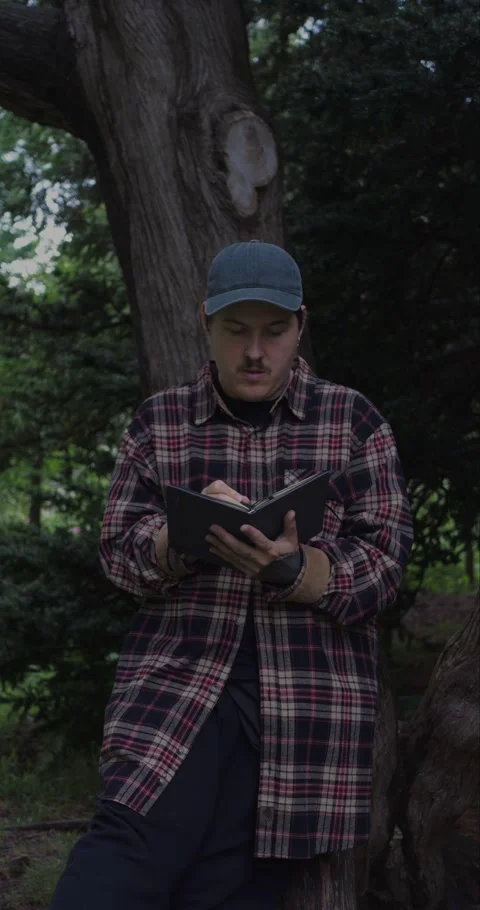Vertical shot of young man wearing a plaid shirt journaling outdoors. Vidéo 313950168
