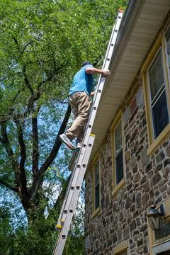 Vertical side view of a man on a ladder cleaning gutters of a stone home Stock Photos