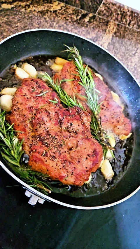 Vertical slow motion of juicy steaks frying with rosemary and garlic in a pan Stock Footage 332480572
