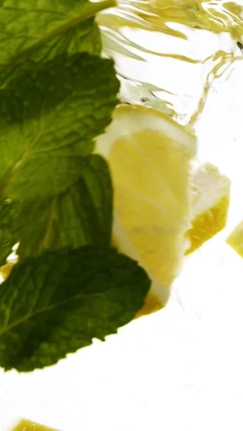 Vertical slow motion slices of lemon fruit and mint leaves splashing in water Stock Footage 268543925