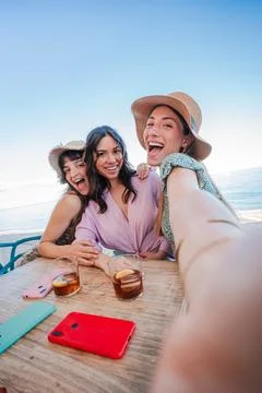 Vertical three girls using a mobile phone to take a selfie, sitting at a beach Stock Photos