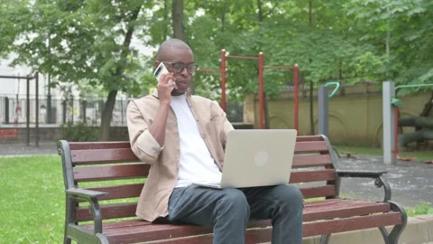 Vertical Video, African Man Talking on Phone While Using Laptop while Sitting Stock Footage 310834019