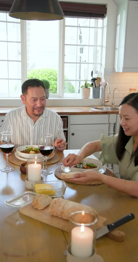 Vertical video: Asian couple smiling at candlelit table slicing bread sipping Stock Footage 345040216