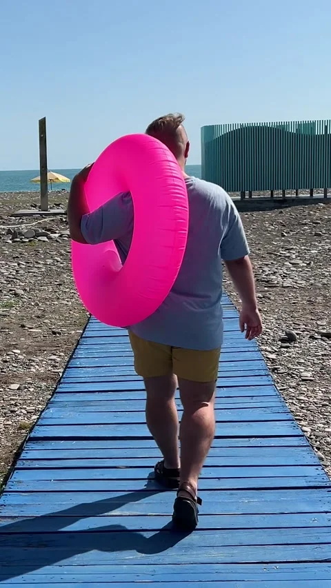 Vertical video. Back view. Man with pink swim ring walks on blue wood path Video stock 331173145