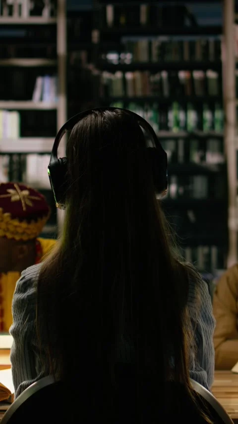 Vertical Video Back view of student adjusting headphones in dark library study Stock Footage 337530149