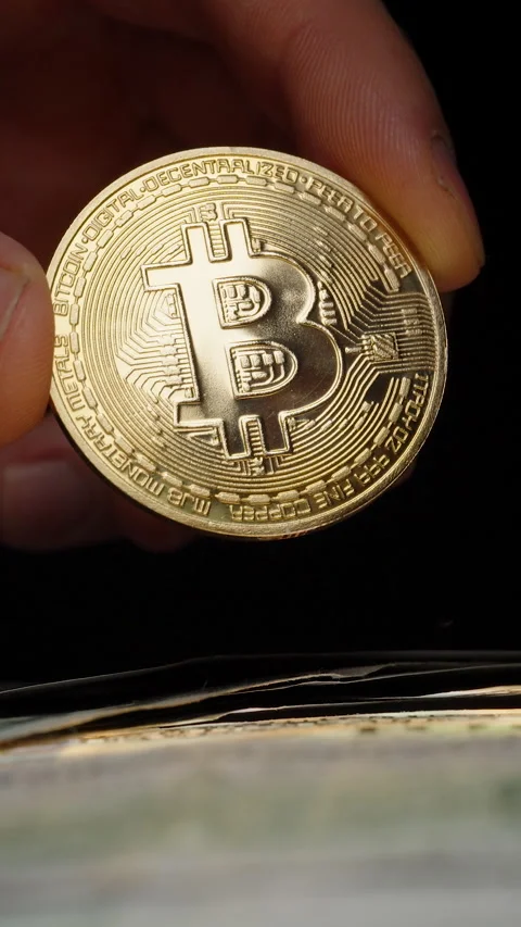 Vertical video. Bare Hand Holding a Shiny Bitcoin Coin Over a Stack of US Dollar Stock Footage 296564935