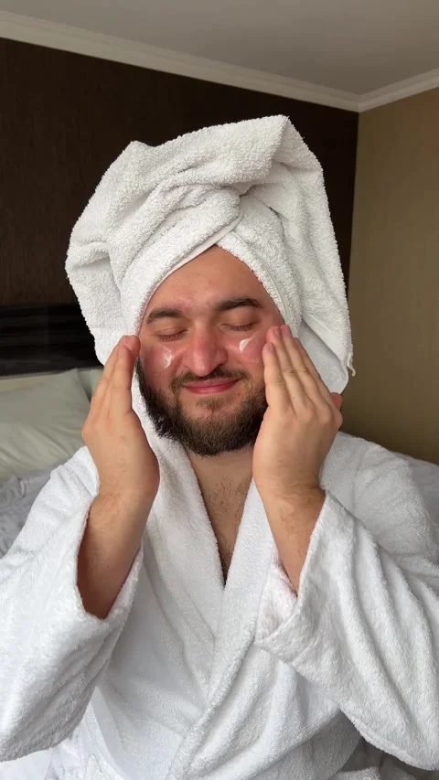 Vertical video. Bearded man in robe, towel on head applies moisturizer to face Stock-Footage 274235966