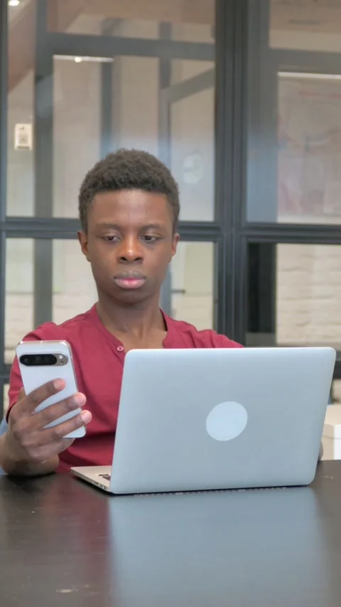 Vertical video of Casual African Man Using Phone with Laptop at Workplace 스톡 동영상 303219476