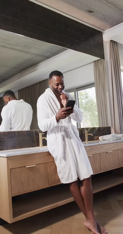Vertical video: Checking phone man in robe smiling brushing with electric Stock Footage 346724156
