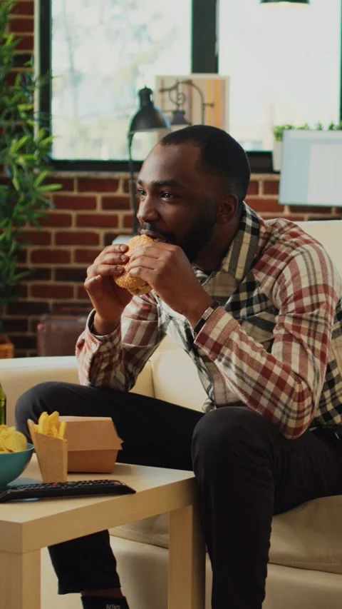 Vertical video: Cheerful man taking bite of hamburger in living room Stock Footage 231852868