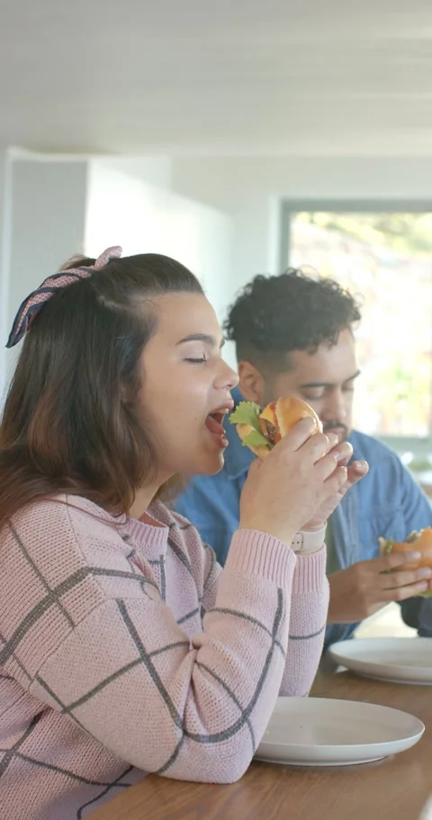 Vertical video: Couple eating plated burgers at kitchen table, woman taking big Stock Footage 345217574