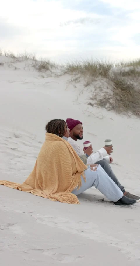 Vertical video: Couple sitting on dune holding cups, man in beanie raising cup Stock Footage 339686373