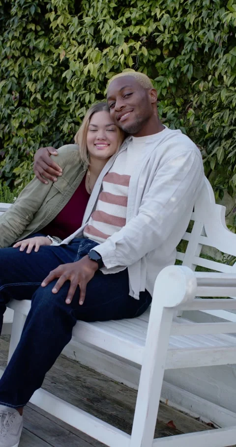 Vertical video: Diverse couple sitting on white bench after camera tilt, leaning Видео 346735355