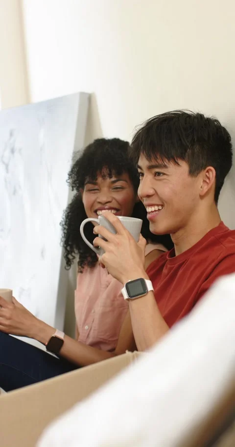 Vertical video: Diverse couple unpacking canvas in living room, sipping from Stock Footage 345040424