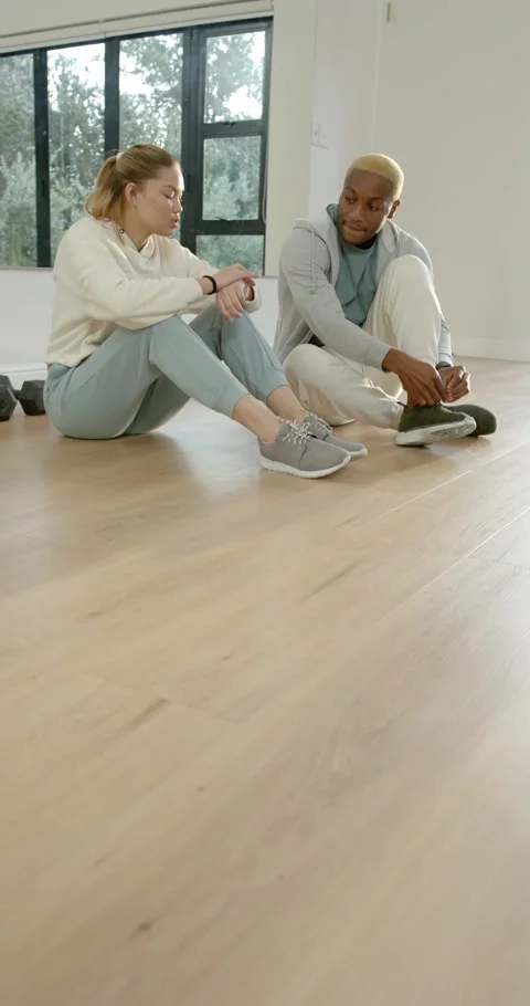 Vertical video: Diverse duo sitting on floor by window, male finishing lacing Видео 346735321