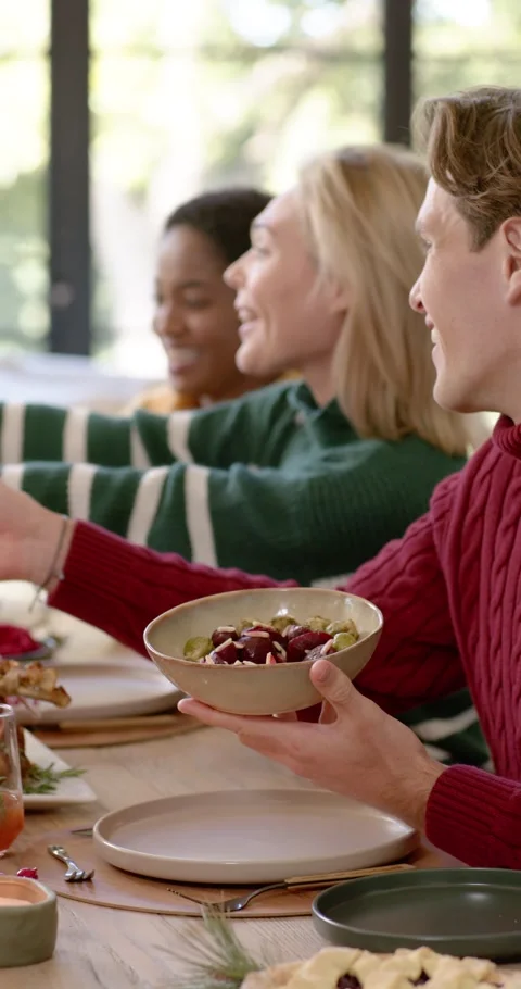 Vertical video: Diverse group sharing at table, man in red knit offering green Видео 346735298