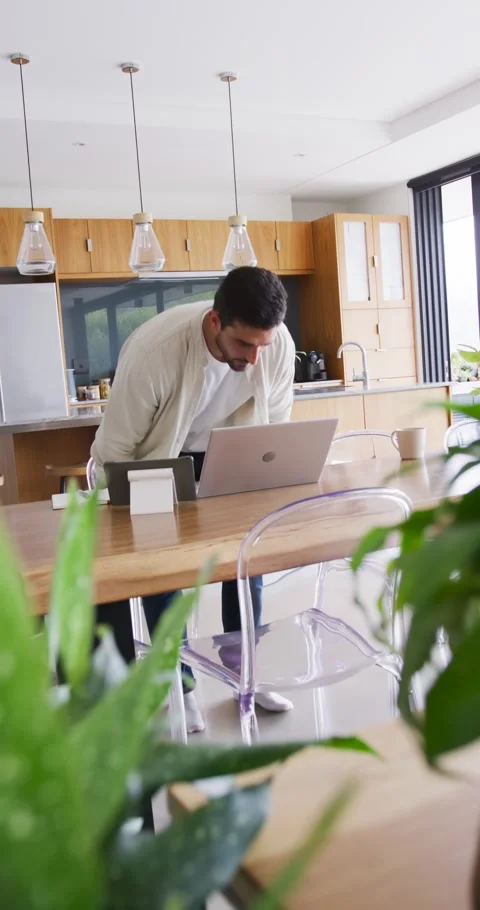 Vertical video: Entering man in white tee arranging laptop at kitchen table, Stock Footage 345397171