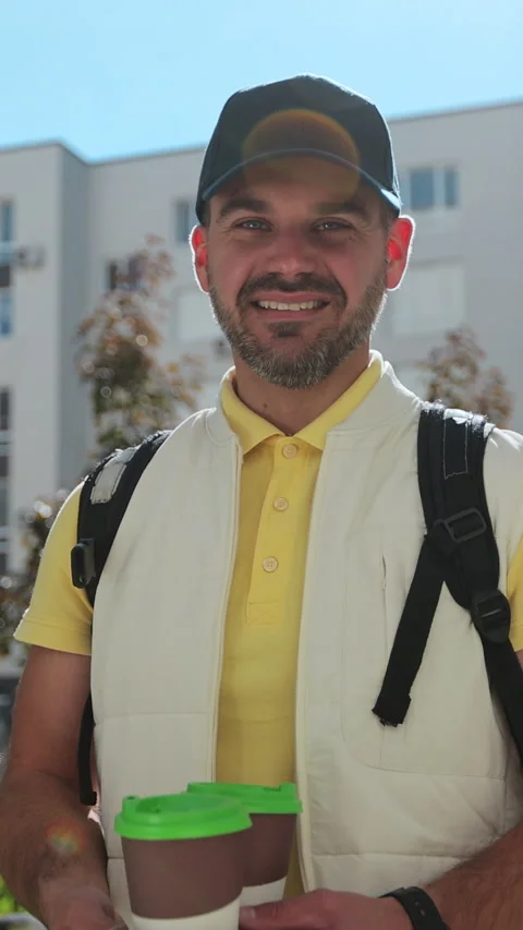 Vertical video. Friendly delivery person in yellow uniform with coffee Видео 344375501